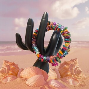 Könnte beinhalten: Ein farbenfrohes Armband mit Edelsteinen, das auf einem schwarzen, handförmigen Ständer präsentiert wird. Das Armband weist eine Vielzahl lebendiger Steine in Rot-, Gelb-, Grün-, Blau- und Lilatönen auf. Muscheln sind auf einer sandigen Oberfläche angeordnet.