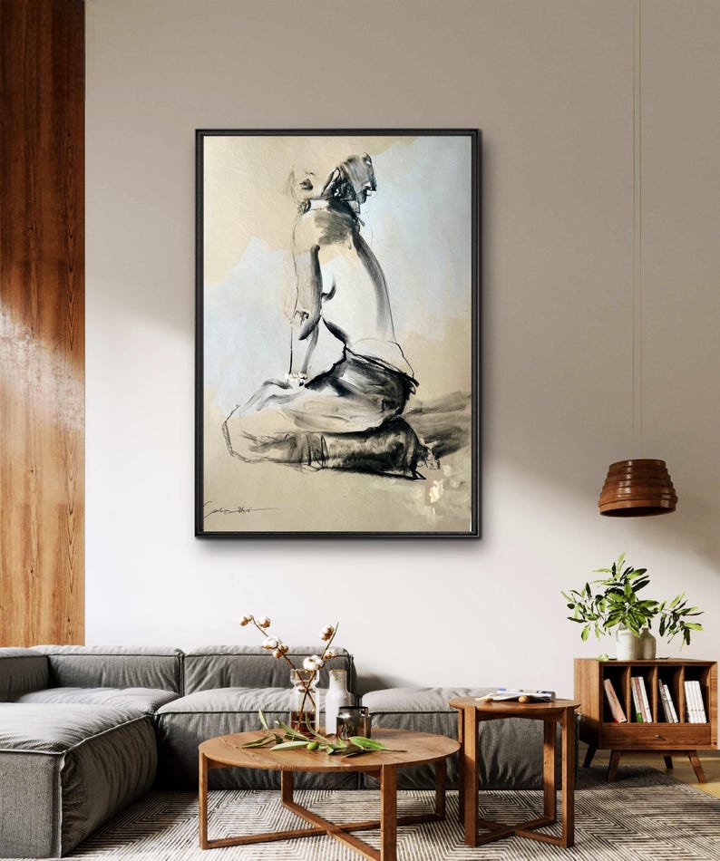 May include: Black and white abstract art print of a seated figure. The artwork is framed in a black frame and hangs on a neutral-colored wall. The art piece is a sketch-like drawing with a minimalist style.