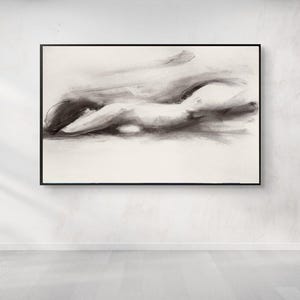 May include: Abstract charcoal drawing of a reclining figure, framed in black. The artwork features soft, blended strokes of gray and black on a cream-colored background. The piece is mounted on a white wall above a light-colored wooden floor.