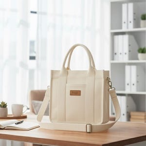 Pode incluir: Bolsa tote creme com formato retangular, com uma alça superior e uma alça de ombro removível. A bolsa tem um pequeno patch retangular com texto na frente. Está em uma mesa de madeira perto de uma janela.