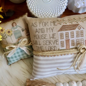 May include: Two decorative cushions with cross-stitch designs. One shows a woman watering sunflowers, the other a house with the text "AS FOR ME AND MY HOUSE WE WILL SERVE THE LORD." Both are tied with twine and bows.