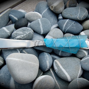 May include: A silver butter knife with a blue glass handle wrapped in wire. The knife is lying on a bed of gray and white stones.