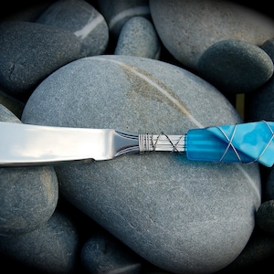 May include: A silver butter knife with a blue glass handle wrapped in wire. The handle is a unique design with a blue and white swirl pattern. The knife is resting on a pile of gray stones.