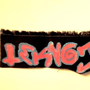 Könnte beinhalten: Ein schwarzer Stoffflicken mit dem Text "LEAKING JUNGLEISM" in roten Buchstaben, die mit hellblau umrandet sind. Der Flicken hat einen ausgefransten Rand und ist vor einem hellgelben Hintergrund platziert.