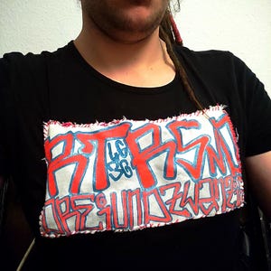 Könnte beinhalten: Schwarzes T-Shirt mit Rundhalsausschnitt und einem großen, rechteckigen Aufnäher mit Graffiti-Text in Rot und Blau. Der Aufnäher ist auf das Shirt genäht. Der Text scheint ein stilisierter Name oder Satz zu sein.