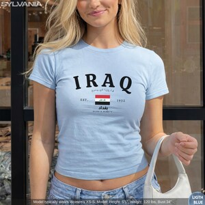 May include: Light blue t-shirt with the word "IRAQ" in black, along with Arabic text and the Iraqi flag. The shirt also features the text "EST. 1932" and the city name "Baghdad".