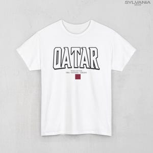 May include: White t-shirt with the word "QATAR" in bold, black outlined letters. Below the word is the text "WORLD CUP 2022 USA-CANADA-MEXICO" and a maroon rectangle. The shirt has short sleeves and a crew neck.