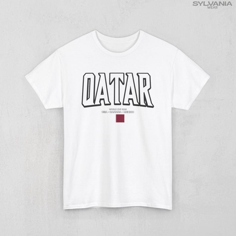 May include: White t-shirt with the word "QATAR" in bold, black outlined letters. Below the word is the text "WORLD CUP 2022 USA-CANADA-MEXICO" and a maroon rectangle. The shirt has short sleeves and a crew neck.