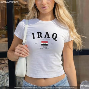 May include: White crew neck t-shirt with the word "IRAQ" in black block letters. Below the word "IRAQ" is the Iraqi flag, the word "Baghdad" in Arabic, and the numbers "1932".