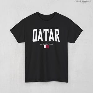 May include: Black t-shirt with the word "QATAR" in large white block letters. Below the word are the words "WORLD CUP 2026" and "USA - CANADA - MEXICO" in smaller white letters. A small flag of Qatar is below the text.