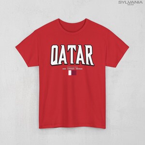 May include: Red t-shirt with the word "QATAR" in large white block letters with a black outline. Below the word "QATAR" is the text "WORLD CUP 2026" and the flags of USA, Canada, and Mexico. The shirt has short sleeves.
