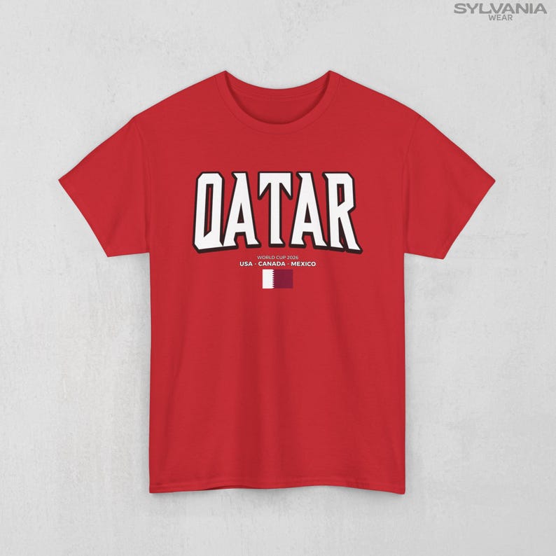 May include: Red t-shirt with the word "QATAR" in large white block letters with a black outline. Below the word "QATAR" is the text "WORLD CUP 2026" and the flags of USA, Canada, and Mexico. The shirt has short sleeves.