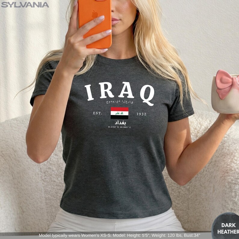 May include: Dark heather gray t-shirt with the word "IRAQ" in white block letters. Below the word "IRAQ" is text in a different language, the Iraqi flag, and the word "Baghdad". The shirt also has the text "EST. 1932".