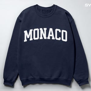 Puede incluir: Sudadera azul marino con la palabra "MONACO" en letras blancas grandes en el pecho. La sudadera tiene cuello redondo y mangas largas. El nombre de la marca "SYLVANIA WEAR" está impreso en blanco en la parte superior derecha.
