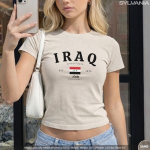 May include: Sand-colored t-shirt with the word "IRAQ" in black, along with Arabic text and the Iraqi flag. The shirt also features the text "EST. 1932" and the city name "بغداد".