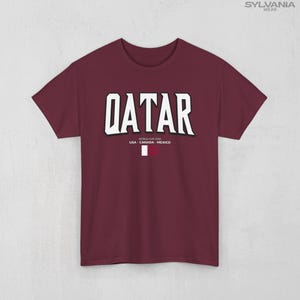 May include: Maroon t-shirt with the word "QATAR" in large white block letters. Below the word are the words "WORLD CUP 2026" and "USA - CANADA - MEXICO". A small Qatar flag is at the bottom. The shirt is from Sylvania Wear.
