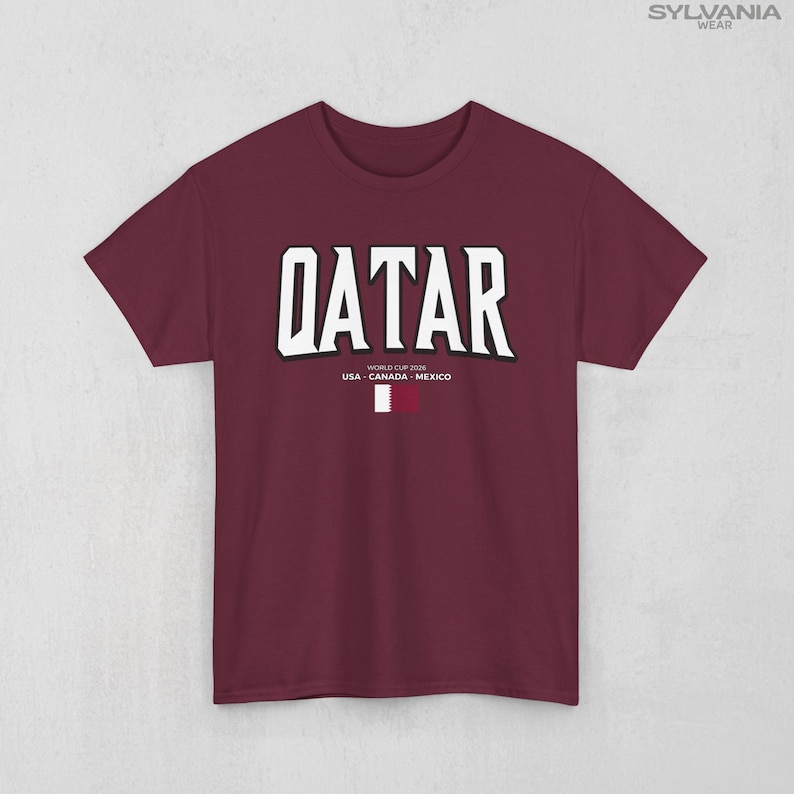 May include: Maroon t-shirt with the word "QATAR" in large white block letters. Below the word are the words "WORLD CUP 2026" and "USA - CANADA - MEXICO". A small Qatar flag is at the bottom. The shirt is from Sylvania Wear.