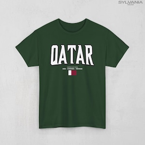 May include: A dark green t-shirt with the word "QATAR" in large white block letters. Below the word are the words "WORLD CUP 2026 USA - CANADA - MEXICO" and a small flag of Qatar.