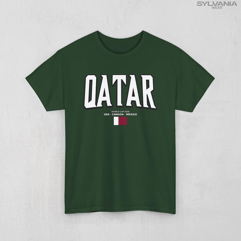 May include: A dark green t-shirt with the word "QATAR" in large white block letters. Below the word are the words "WORLD CUP 2026 USA - CANADA - MEXICO" and a small flag of Qatar.
