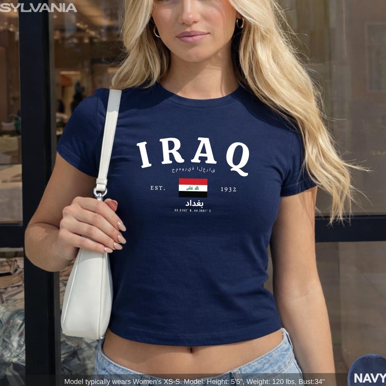 May include: Navy blue t-shirt with the word "IRAQ" in white block letters. Below the word "IRAQ" is the text "EST. 1932" and the Iraqi flag. The shirt also has Arabic text and the word "Baghdad".