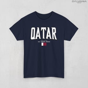 May include: Navy blue t-shirt with the word "QATAR" in large white block letters. Below the word "QATAR" is the text "WORLD CUP 2026 USA - CANADA - MEXICO" and a small flag of Qatar.