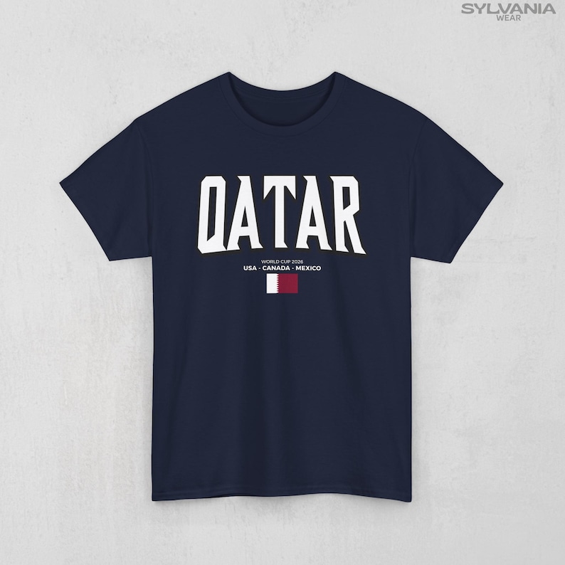 May include: Navy blue t-shirt with the word "QATAR" in large white block letters. Below the word "QATAR" is the text "WORLD CUP 2026 USA - CANADA - MEXICO" and a small flag of Qatar.