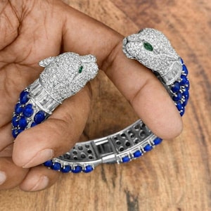 Peut inclure: Un bracelet en argent orné de deux têtes de panthère serties de cristaux transparents et d'yeux verts. Le bracelet est orné de pierres bleues. Le bracelet est présenté sur une surface en bois.