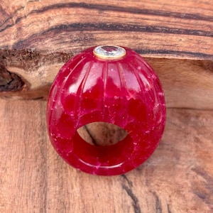 May include: A bright red ring with a carved, ribbed pattern. The ring has a clear, oval gemstone set in a gold-coloured bezel. The ring is displayed on a wooden surface.