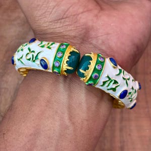 以下が含まれることがあります： 白いエナメルベースに緑の花柄が施され、ゴールドトーンの金属縁で装飾されたヒンジ付きバングルブレスレット。中央のヒンジ付近の緑色のバンドには、楕円形の青い石と小さな円形の紫色の石が埋め込まれています。