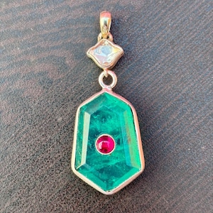 Puede incluir: Colgante dorado con una piedra verde talla esmeralda y una pequeña piedra roja en el centro. El colgante tiene una piedra transparente en forma de diamante en la parte superior y una fianza dorada. El colgante mide unos 5 cm de largo.