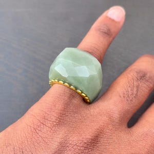 以下が含まれることがあります： ファセットデザインの、大きくて明るい緑色の宝石リング。 リングは、スカラップエッジのゴールドカラーのバンドが付いています。 リングは指に着用されています。