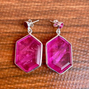 May include: A pair of silver-toned earrings with a geometric design. Each earring features a large, faceted, magenta-colored gemstone set within a silver frame. A smaller, round, magenta stone tops each earring.