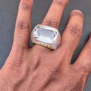 以下が含まれることがあります： 透明な長方形のファセットカットストーンをあしらった大ぶりのリング。半透明の明るい色の樹脂製バンドにセットされています。セッティングのベース部分には、指を囲むように細い質感のあるゴールドトーンの金属トリムが施されています。