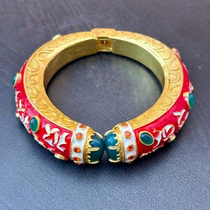 以下が含まれることがあります： 鮮やかな赤のエナメル表面に白い花柄が施された、ゴールドトーンのヒンジ付きバングルブレスレット。縁と留め具部分には、楕円形の緑色のカボションストーンと小さな丸いオレンジ色のクリスタルがあしらわれています。