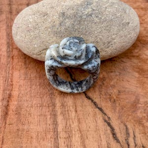 May include: A carved ring featuring a rose design. The ring is made of a speckled white and gray stone. The ring is set against a wooden surface and a large, smooth stone.