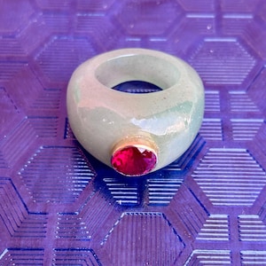 Peut inclure: Une bague vert clair avec une grosse pierre rouge ovale sertie dans une monture dorée. La bague a un design lisse et arrondi, sur un fond violet.