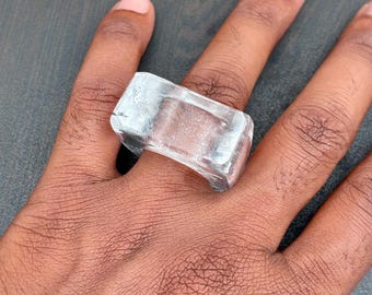 May include: A clear, rectangular ring is worn on a finger. The ring appears to be made of a transparent material, possibly glass or crystal, with a slightly rounded band. The ring's shape is geometric and modern.