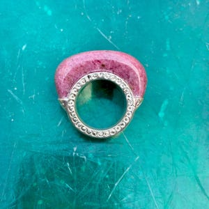 Puede incluir: Un anillo con una gran gema rosa ovalada engastada en una banda plateada. La banda está adornada con pequeñas piedras transparentes. El anillo está sobre un fondo turquesa.