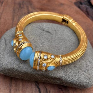 May include: A gold-toned hinged cuff bracelet featuring a ribbed band and two ornate terminals. Each terminal is set with a smooth, round, light blue cabochon stone, accented by small, clear, sparkling round-cut stones in a decorative setting.