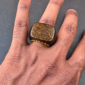 以下が含まれることがあります： ダークブラウンの木製バンドと、金茶色と黒のまだら模様の有機的なパターンが施された平らで磨かれた上面を持つ長方形のシグネットリング。リングバンドの裏側には、明るい金色の金属製の裏地が見えます。