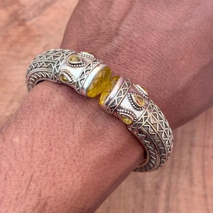 Op de afbeelding: Zilverkleurige armband met ingewikkelde geometrische patronen en gele edelsteenaccenten. De armband heeft een centrale gele edelsteen en kleinere gele stenen in decoratieve zilveren zettingen. Een uniek sieraad.
