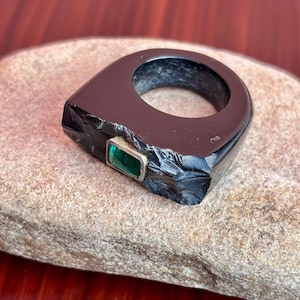 May include: A black ring with a rectangular, emerald-green stone set in a silver-colored metal. The ring has a unique, angular design and sits on a light brown, textured surface.