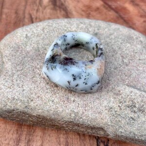 May include: A thick, faceted ring carved from white stone featuring dark, fern-like dendritic inclusions throughout its surface. The band has a smooth, polished finish with a rounded, chunky profile and a distinct natural pattern of black branching marks.