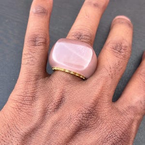 以下が含まれることがあります： 角が丸みを帯びた大きな淡いピンク色の長方形の石が特徴のチャンキーリング。石は指を囲む細い質感のあるゴールドトーンの金属バンドの上にセットされており、柔らかなピンクとメタリックゴールドのコントラストを生み出しています。