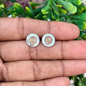 Op de afbeelding: Een paar ronde oorstekers. Elke oorbel heeft een licht perzikgekleurde edelsteen gezet in een zilverkleurige bezel. De oorbellen worden getoond op de hand van een persoon, met groene bladeren op de achtergrond.