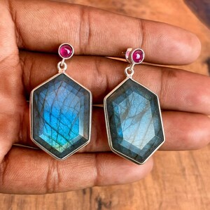 May include: A pair of silver earrings featuring large, faceted, hexagonal labradorite stones with blue and green iridescence. Each earring has a small, round, red gemstone at the top.