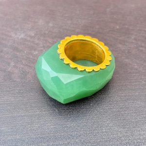 以下が含まれることがあります： ゴールドトーンのメタルバンドにセットされた、大きなファセットカットのライトグリーンの石が特徴のリング。バンドにはリングの開口部を縁取る装飾的なスカラップエッジがあり、グリーンの石の滑らかで角張った表面と対照的です。