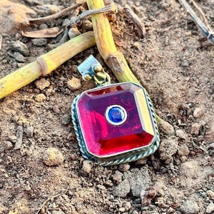 May include: A square pendant featuring a large, faceted, translucent red stone with a smaller, circular, deep blue stone set in the center. The pendant is framed by a twisted silver-toned metal border and includes a decorative silver-toned bail.