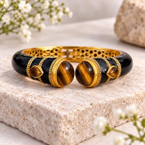 May include: A hinged cuff bracelet featuring a polished black enamel finish and gold-tone metal accents. The design includes two large, round, brown tiger's eye cabochons and two smaller, teardrop-shaped tiger's eye stones at the open ends.