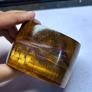 Könnte beinhalten: Ein poliertes Armband aus Tigeraugenstein. Das Armband zeigt Bänder in Goldgelb, Braun und Rotbraun. Der Stein hat eine glatte, gebogene Form und eine glänzende Oberfläche, die die natürlichen Muster zeigt.